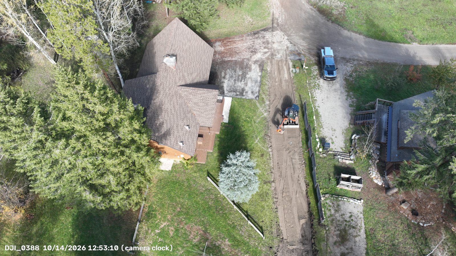 Low angled drone photo showing a compact excavator grading a dirt strip between two houses, with a fence along its east edge